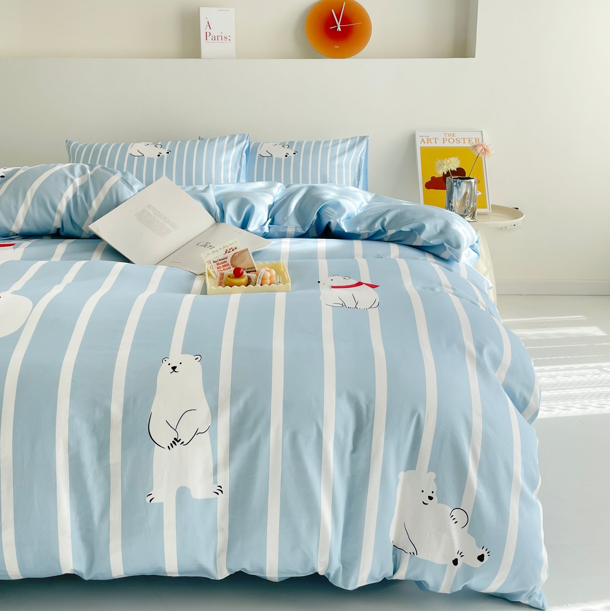 A bright and cozy bedroom featuring a cute bedding set with a light blue and white striped Polo Bear bed comforter. The adorable polar bear illustrations add a playful touch, while matching pillows, an open book, and a small tray of desserts create a relaxing and inviting atmosphere.