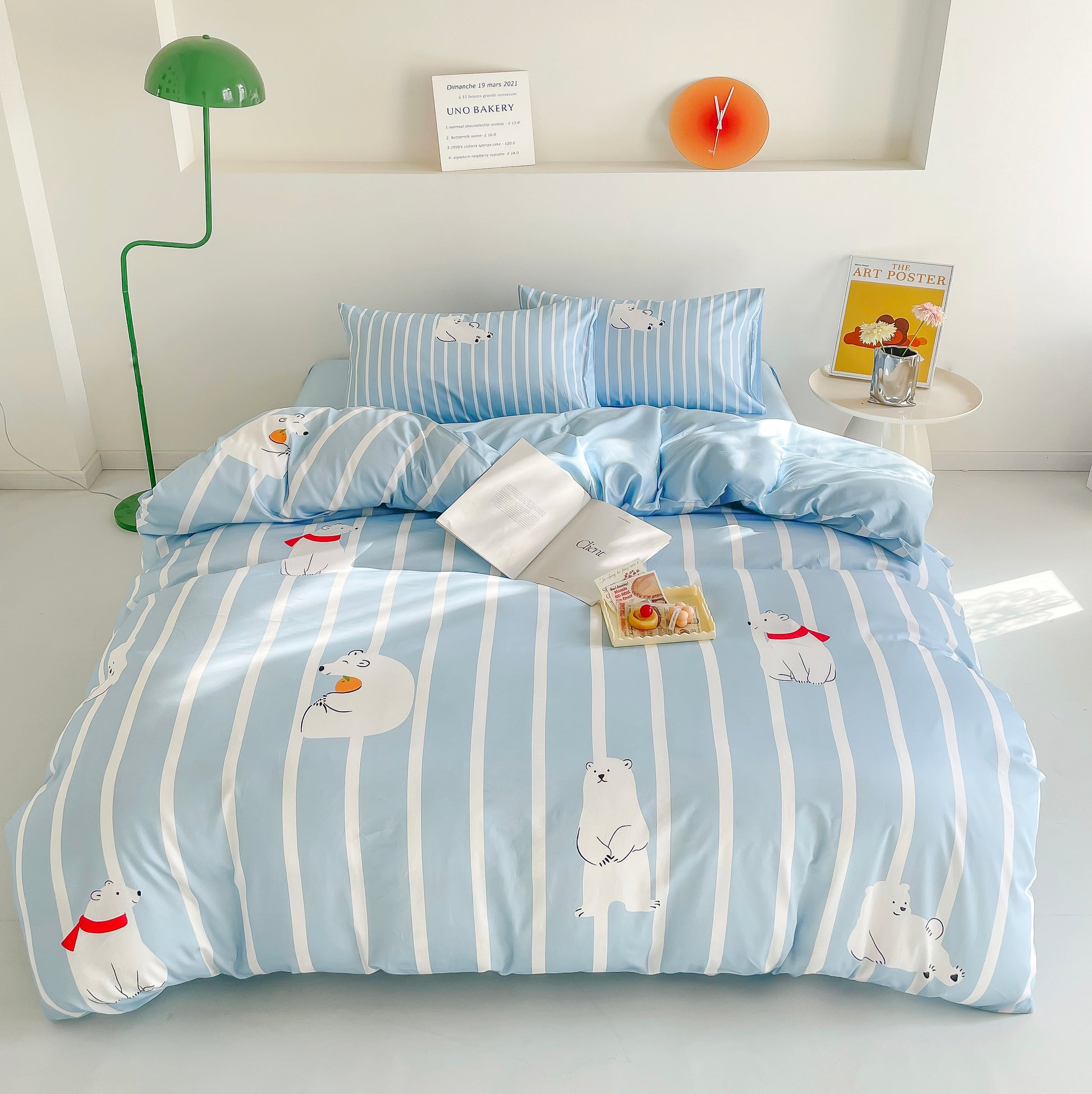 A bright and cozy bedroom featuring a light blue and white striped bedding set with adorable polar bear illustrations. The bed is neatly arranged with matching pillows, an open book, and a small tray of desserts, creating a relaxing and inviting atmosphere.